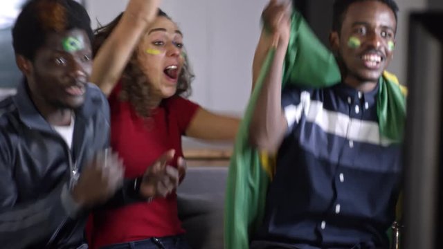 Two Brazil men and young woman with flag and face paint supporting their team, shouting in excitement, raising arms and embracing while watching soccer on TV at home