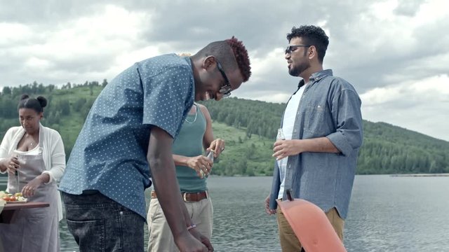 Group of six diverse people preparing food for picnic, talking and having fun, men grilling meat and women cutting vegetables at table