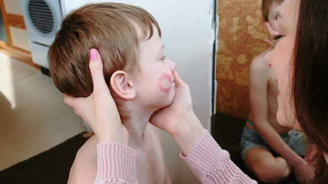 Mom kisses her son on the cheek and leaves a trace of lipstick. They're sitting in front of the mirror.