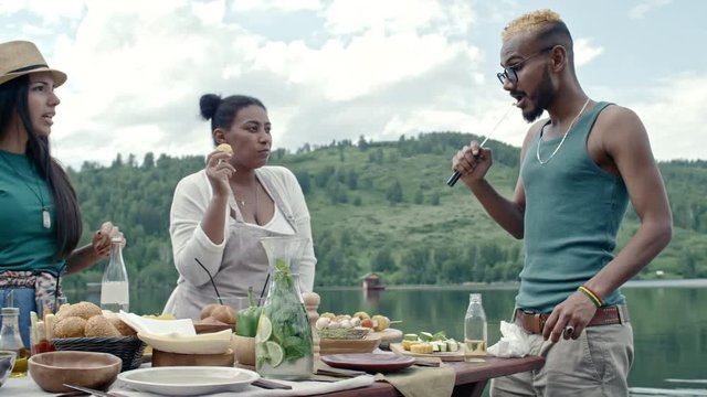Three friends of different ethnicities standing at table and enjoying food at picnic, man trying something off his knife