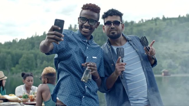 Two black men standing together and taking selfie on smartphone, one holding drinks and one holding barbecue tongs