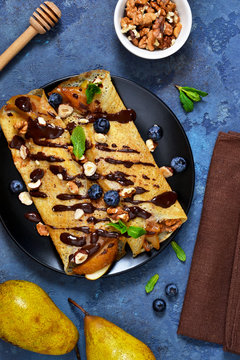 Homemade thin pancakes with caramelized pears, chocolate sauce and nuts on a concrete background.