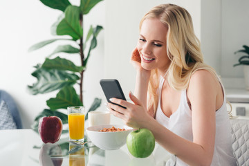 beautiful smiling blonde girl using smartphone while having breakfast at home