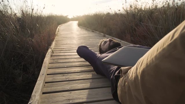 Dreaming adult man relaxing from daily rush lying on wooden walkway in field with laptop near.