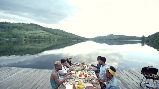 Side view of six young friends of diverse ethnicities sitting at table on wooden dock and enjoying picnic by lake and laughing