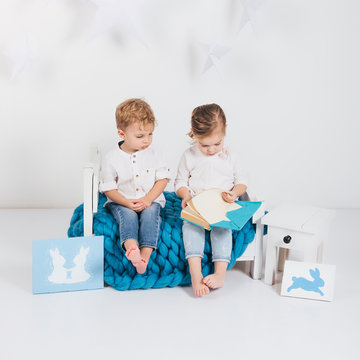 Adorable Little Kids Sitting On Blue Knitted Plaid And Reading Book And Rabbit Cards, Easter Concept