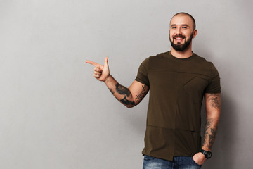 Image of unshaved joyful guy with tattoos on hands posing at camera and pointing finger aside on...