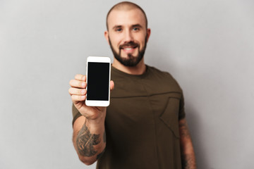 Portrait of smiling muscular man 30s with beard showing smartphone with black screen on camera, isolated over gray background