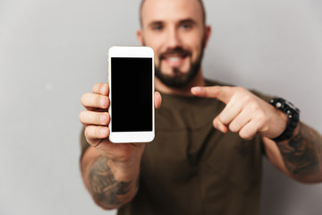 Photo of blurry man 30s holding mobile phone and pointing finger on black empty screen in focus, isolated over gray background