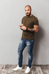 Full-length image of muscular man 30s looking on camera while using smartphone holding in hand, isolated over gray background