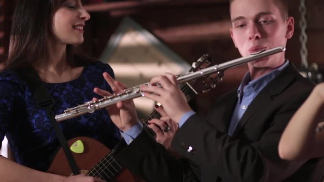 A guy in a jacket and shirt plays the flute, and a beautiful brunette in a blue dress playing the violin. Two people musician reproduce music.