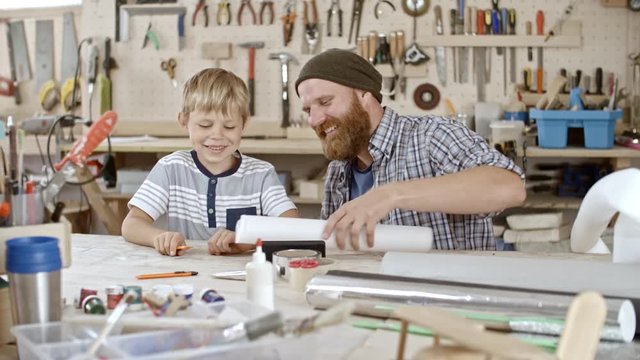 Father and son sitting in art studio, making crafts and smiling when carving something out of Styrofoam roll