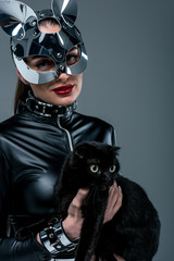 Kinky woman in sexy costume and mask holding black cat isolated on grey