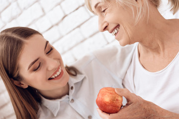 Girl is nursing elderly woman at home. They are embracing. Woman is holding apple.