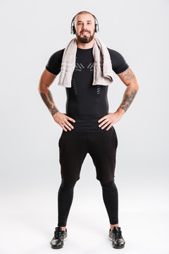 Full-length photo of muscular man listening to music via headphones while standing with hands on waist and towel on neck after sport, isolated over white background