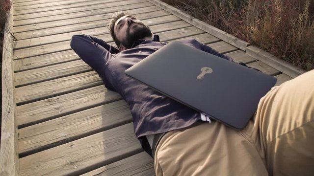 Dreaming adult man relaxing from daily rush lying on wooden walkway in field with laptop near.