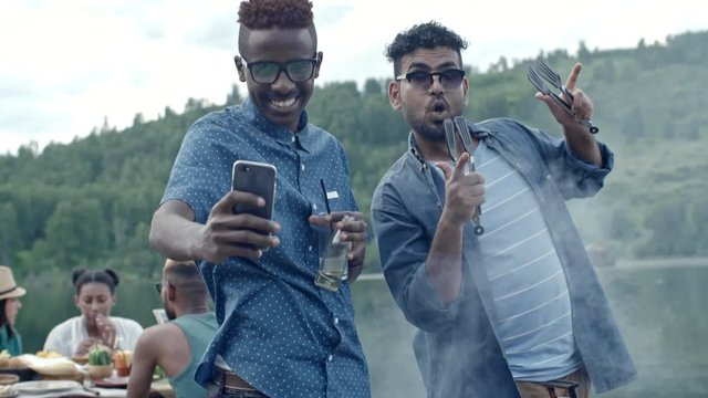 Two black men taking selfie together and posing for phone camera, one holding drinks and one holding barbecue tongs