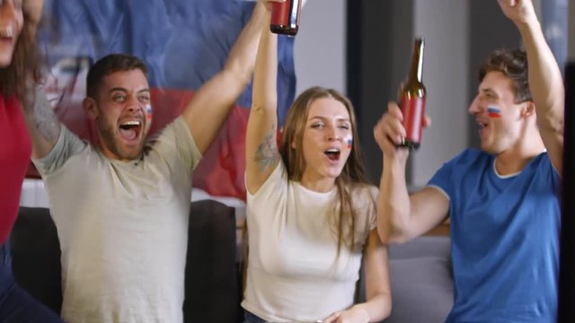 Group of young cheerful Russian soccer fans with flag paint on their cheeks sitting on couch and watching TV game. Man screaming and shaking with flag of Russia while friends toasting with beer