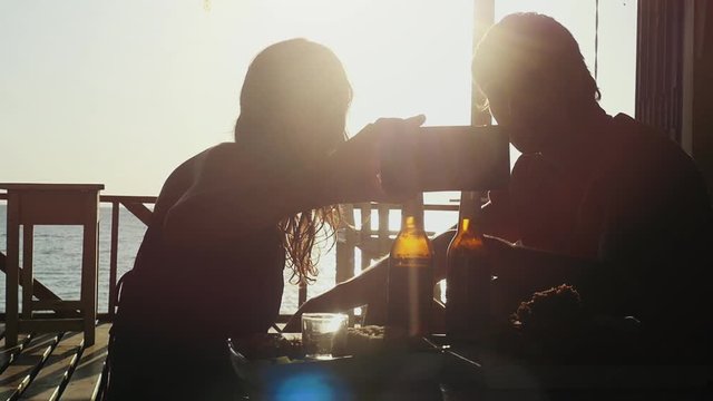 Young beautiful couple takes selfie with smartphone having rest in outdoor beach cafe drinking beer at amazing sunset time in slow motion. 1920x1080