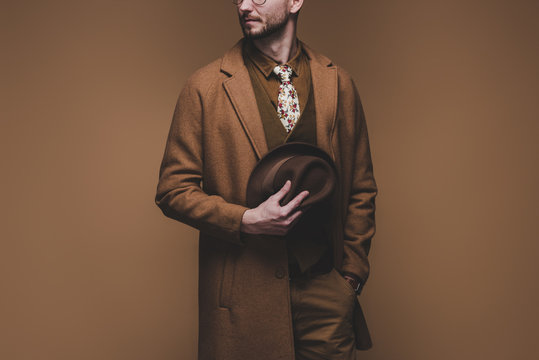 Stylish Man Wearing Coat And Holding Hat Isolated On Brown