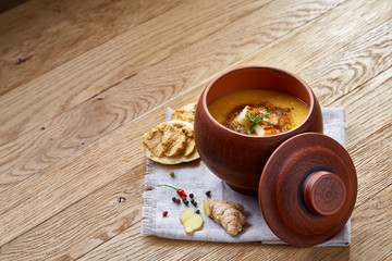 Clay pot of pumpkin soup on homespun napkin rustic wooden background, close-up, selective focus, top view.