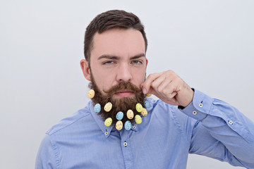 Portrait of Handsome bearded man looking at camera and holding his mustache with Easter decoration in his beard on white background. Happy Easter greeting card concept © Kira_Yan