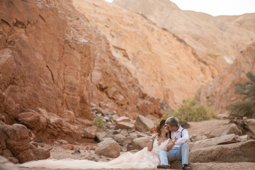 Bride and groom sit and kiss in canyon on background of rocks.