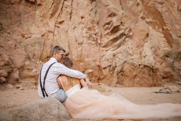 Bride and groom sit and embrace on sand in canyon. Back view.