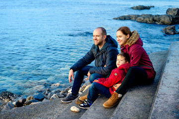 Fototapeta premium family mom, dad and son are sitting on the beach by the sea and looking into the distance. Season - spring, autumn. The concept of family happiness