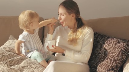 Little girl eating yogurt and feeding mother sitting on sofa