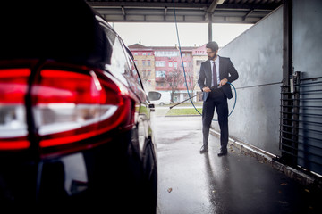 Rear view of the car and elegant stylish young focused man in a suit washing tire of the car with a...