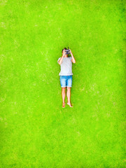 A man lies on a green lawn and smiles with happiness. Top and aerial view with copy space. Minimal styled flat lay isolated on original green grass background. Looks at the phone lying on his back.