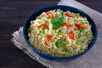 Instant noodles on top with chili spring onion and cilantro in dark blue bowl on a sack in wooden background