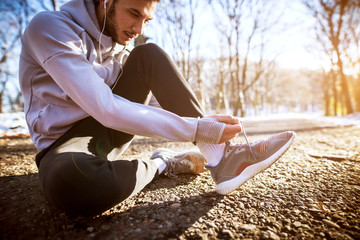 Handsome young man tying shoelaces in the winter forest while sitting on the ground.