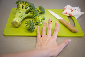 Wounded finger with blood and knife on cutting board with broccoli on grey background