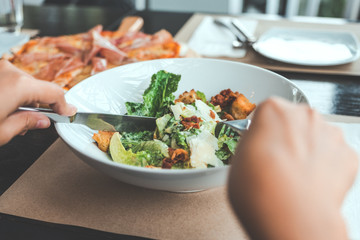 A hand using fork and knife to eat Ceasar salad with pizza on dining table in the restaurant