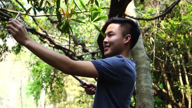 Selfie photo concept : Asian young man holding smartphone for selfies at  forest, beautiful scenery for peaceful in spring , It shows moisture, tranquility and refreshing of rain forest in Thailand