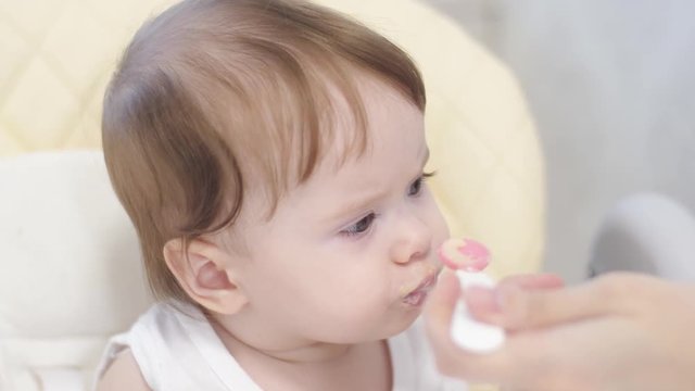 Baby eats porridge from spoon, spits and smiles sitting on highchair in kitchen. First feeding of babies. Play.