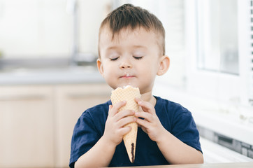 A child in a dark-blue t-shirt in the bright kitchen eating a waffle ice cream cone in the summer house