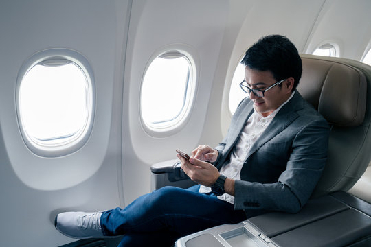 Young Asian Businessman With Suit Sitting In Business First Class Seat Via Using Smartphone With Happiness