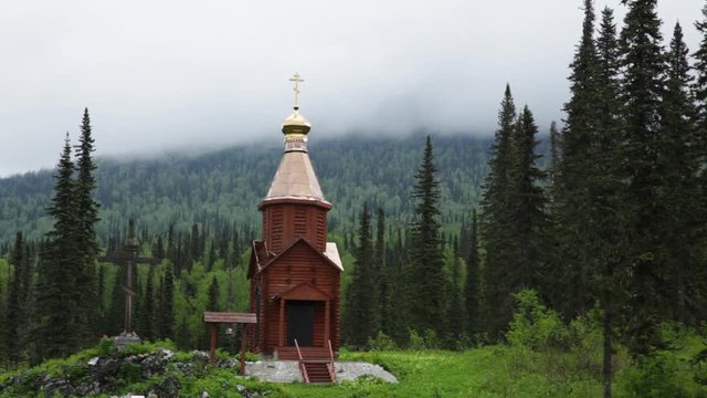 Chapel with a dome and a cross in the mountains of Khakassia against a background of tall green spruce trees. Mountain peaks covered in blue fog. Travel video without people.