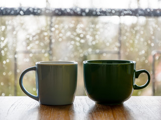 Two mugs on a wooden table in front of a glass window covered with rain drops.