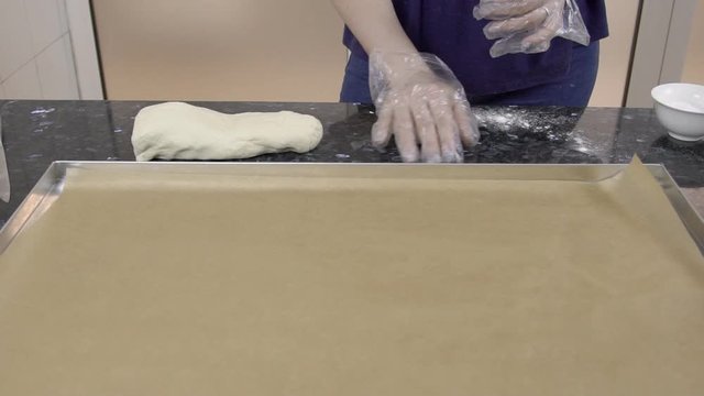 Female baker is rolling small piece of dough on black table at kitchen. Big baking sheet is lying on the desk and covered with brown paper. Lady in blue blouse and gloves and putting future pies to