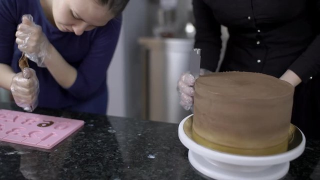 Two confectioners work while standing at kitchen table in pastry shop. Two women make chocolate figures and level surface of chocolate cake in cuisine indoors. Female professionals are in working