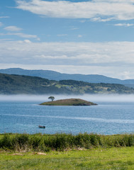 Green islet in a blue bay in a fog on a background of mountains, Norway