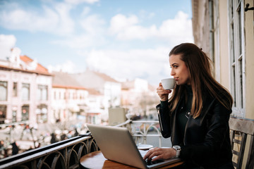 Woman drinking coffee on the terrace wihle working on a laptop.
