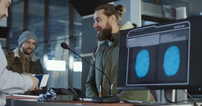 Airport security checking a thumb print with a close up view of a computer screen processing the match while two male passengers standing in a queue at the counter behind.