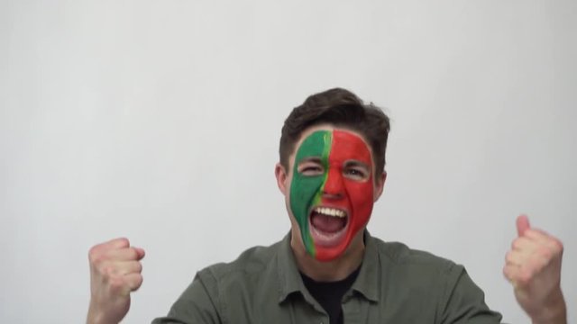 Handsome man supporter fan of Portugal national team painted flag face get happy victory screaming into a camera. Football emotions.