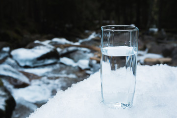 A transparent glass glass with drinking mountain water stands in the snow against a background of a...