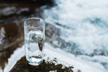 A transparent glass glass with drinking mountain water stands in the moss stone on sun beame against a background of a clean frost mountain river. The concept of drinking mountain drinking mineral